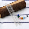 A bundle of stainless steel beading needles is laid across a wooden cylinder on a white cloth.