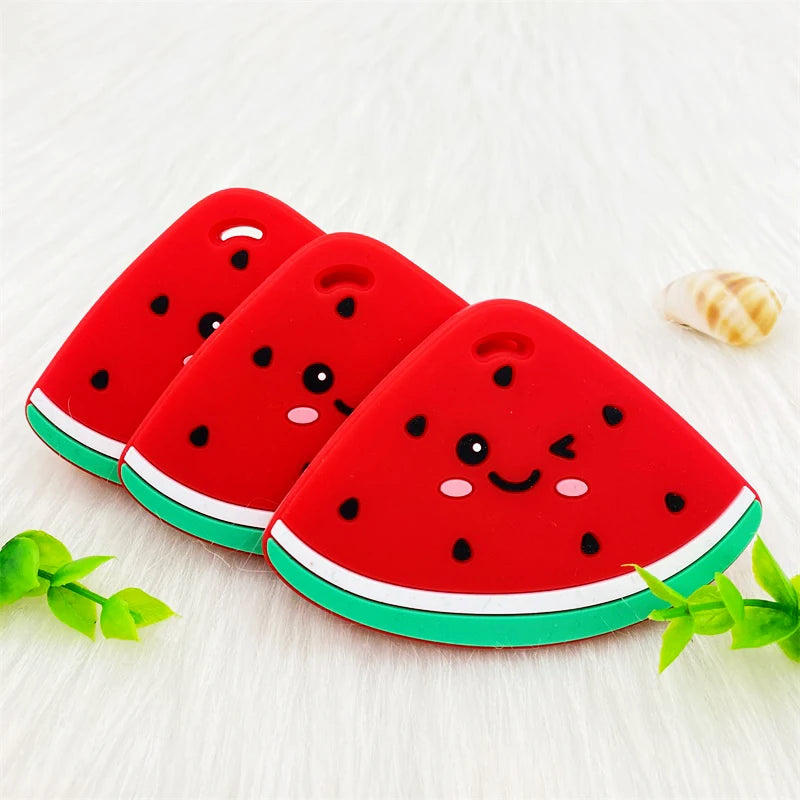 Red watermelon silicone teether pendant with a smiling face resting on a white surface.