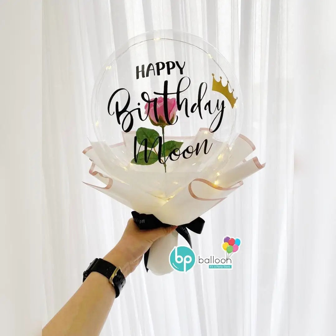 A hand holds a clear balloon featuring the text Happy Birthday Moon with a gold crown and green leaf.