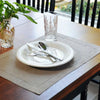 A place setting on a wooden table featuring a white plate, silverware, and a glass, set against a neutral linen napkin.