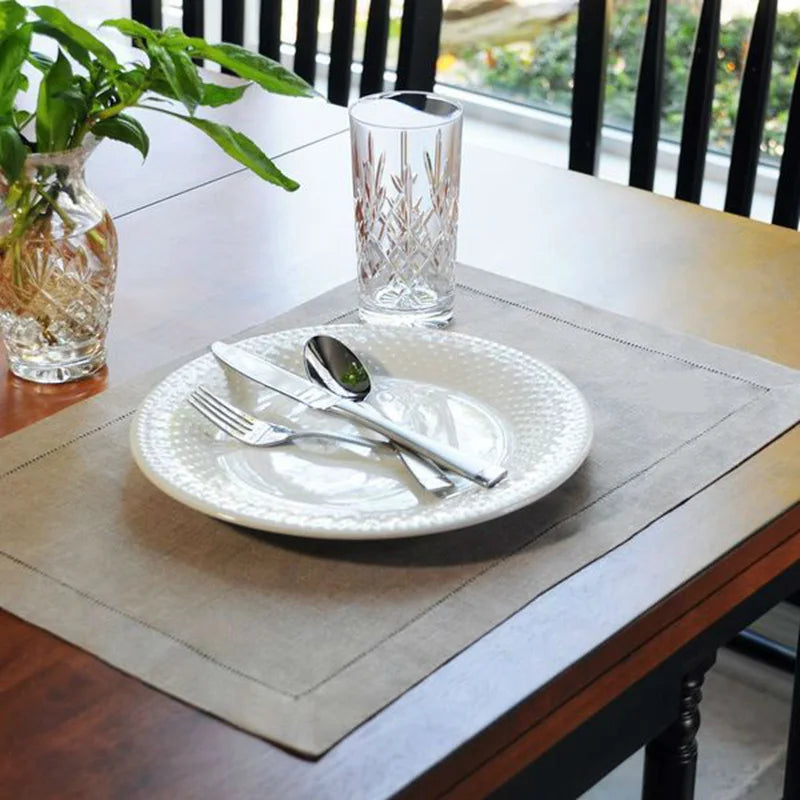 A place setting on a wooden table featuring a white plate, silverware, and a glass, set against a neutral linen napkin.