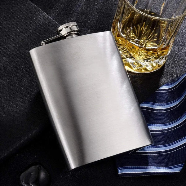 A silver stainless steel hip flask with a metal funnel is shown next to a glass of amber liquid and a striped cloth.