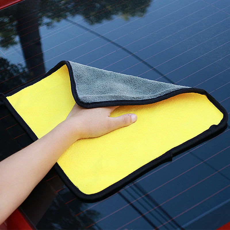 A hand is holding a yellow microfiber car care towel with a gray trim against a dark blue car surface.