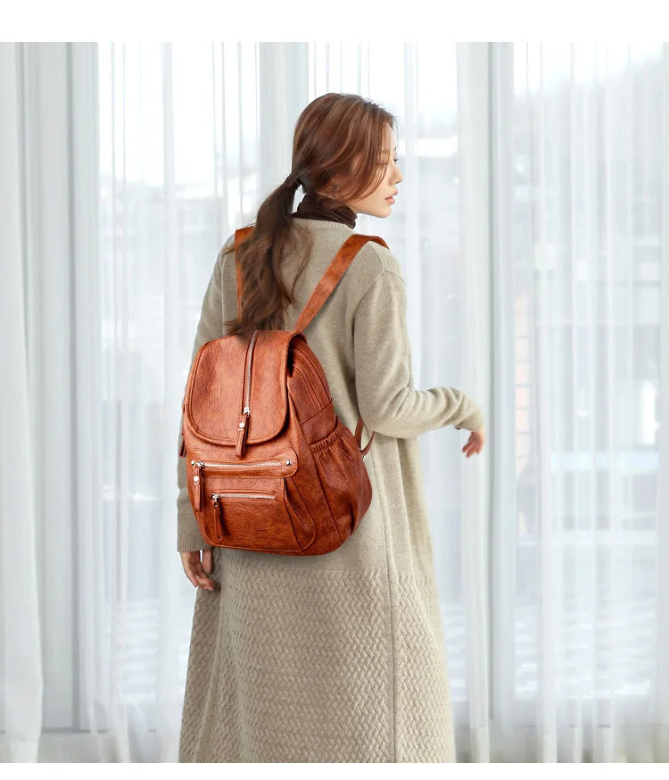 A woman wearing a tan coat carries a brown vintage faux leather backpack with multiple compartments.