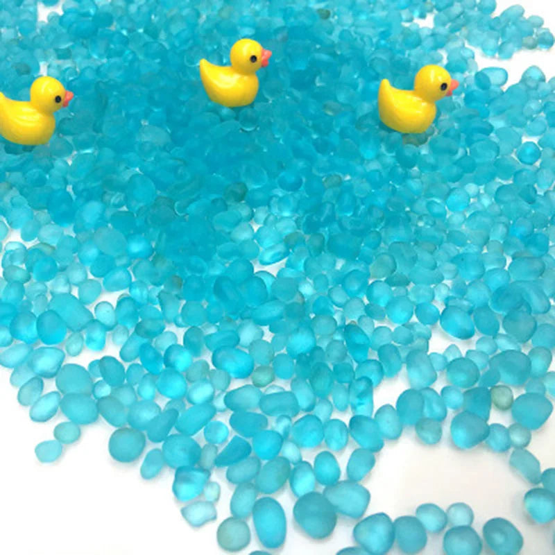 A pile of bright blue decorative stone pebbles with small yellow duck figurines scattered across a white surface.