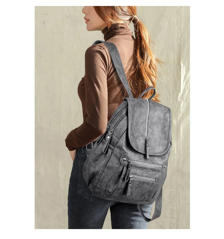 A woman wearing a brown turtleneck and dark jeans is carrying a gray vintage faux leather backpack.