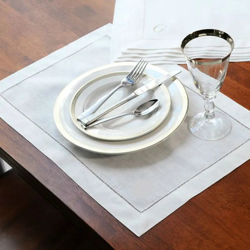 A set of six linen hemstitch napkins, silverware, and a glass are displayed on a wooden table.