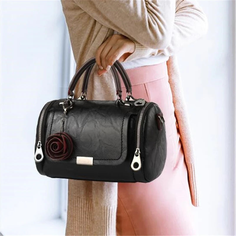 A woman wearing a beige sweater holds a black leather crossbody bag with a floral pendant.