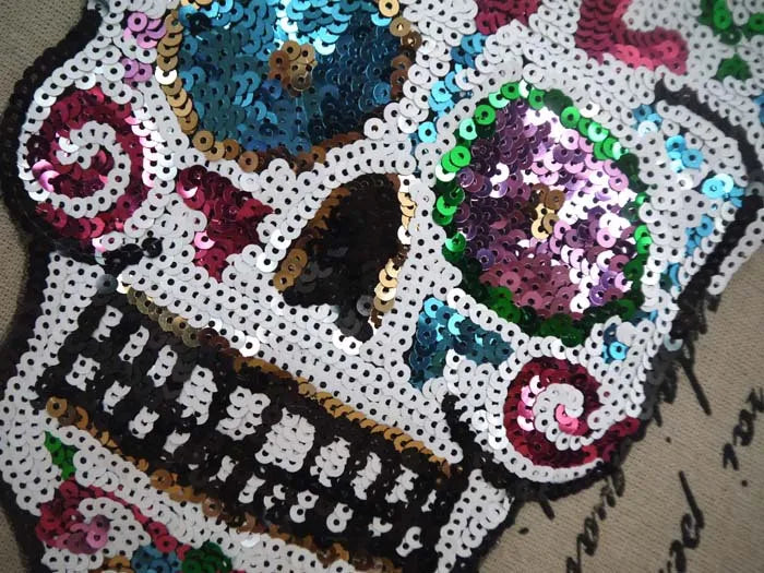 A close-up of a sequin skull embroidered patch featuring purple, teal, and pink sequins on a white background.
