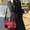 A woman holds a red leather crossbody bag with a floral pendant against a blurred background.