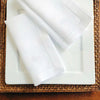 A set of six white linen hemstitch napkins is displayed on a white plate atop a woven mat.