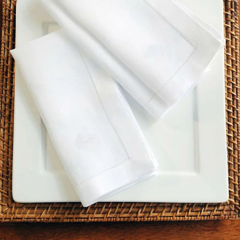 A set of six white linen hemstitch napkins is displayed on a white plate atop a woven mat.