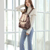 A woman wearing a beige turtleneck sweater and blue pants stands in front of a window, carrying a brown faux leather backpack