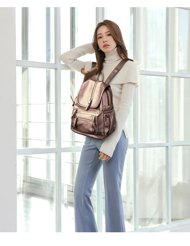 A woman wearing a beige turtleneck sweater and blue pants stands in front of a window, carrying a brown faux leather backpack