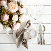 A flat lay of a linen hemstitch napkin set with six folded napkins, silverware, and a glass on a white wooden table.