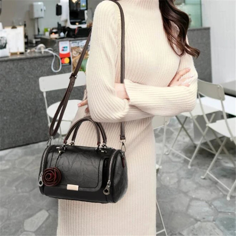 A woman wearing a cream knit dress holds a black leather crossbody bag with a floral pendant.
