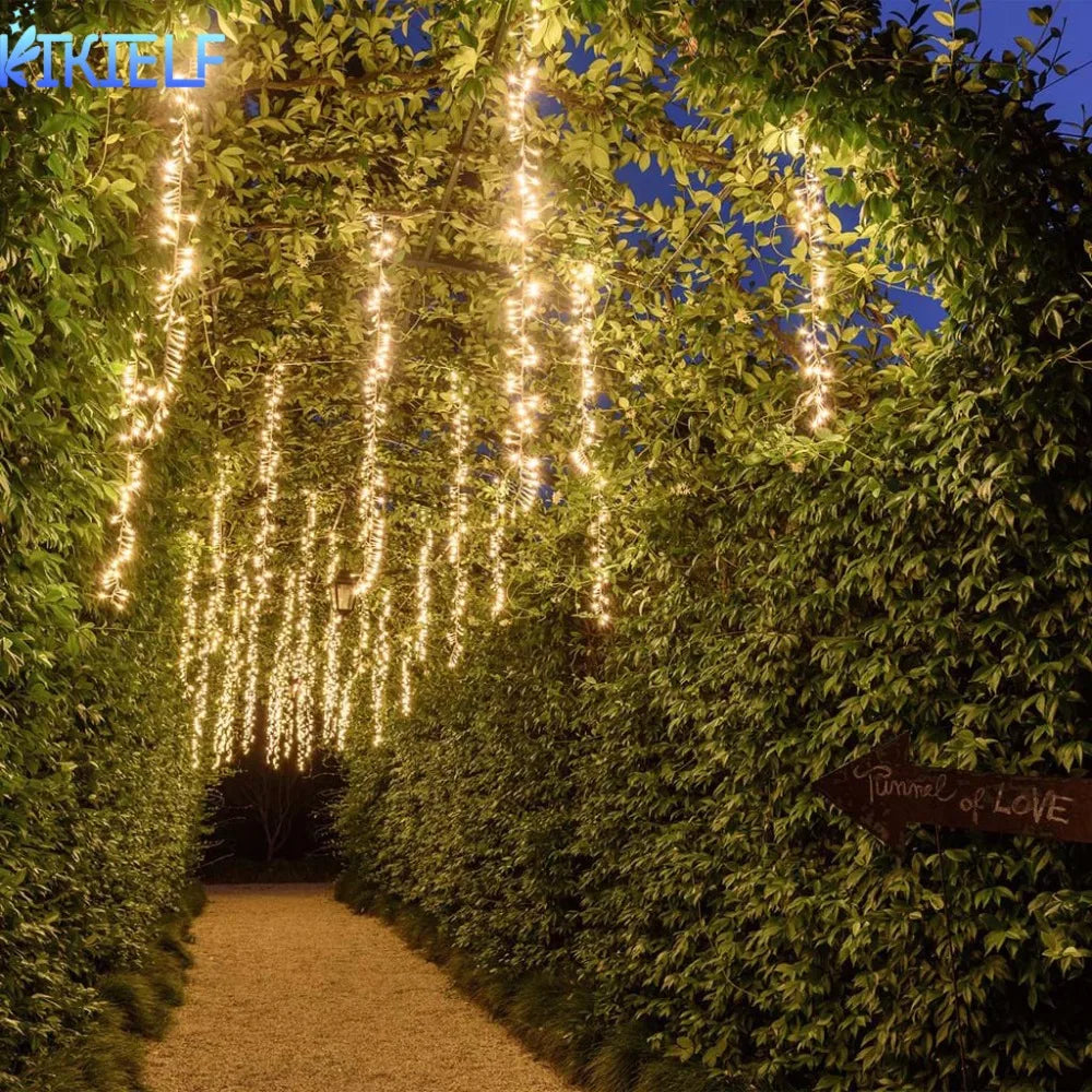 A lush green hedge tunnel illuminated by vertical fairy lights leading into the distance.