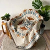 A bohemian jacquard woven throw blanket draped over a wooden chair next to a large green monstera plant.