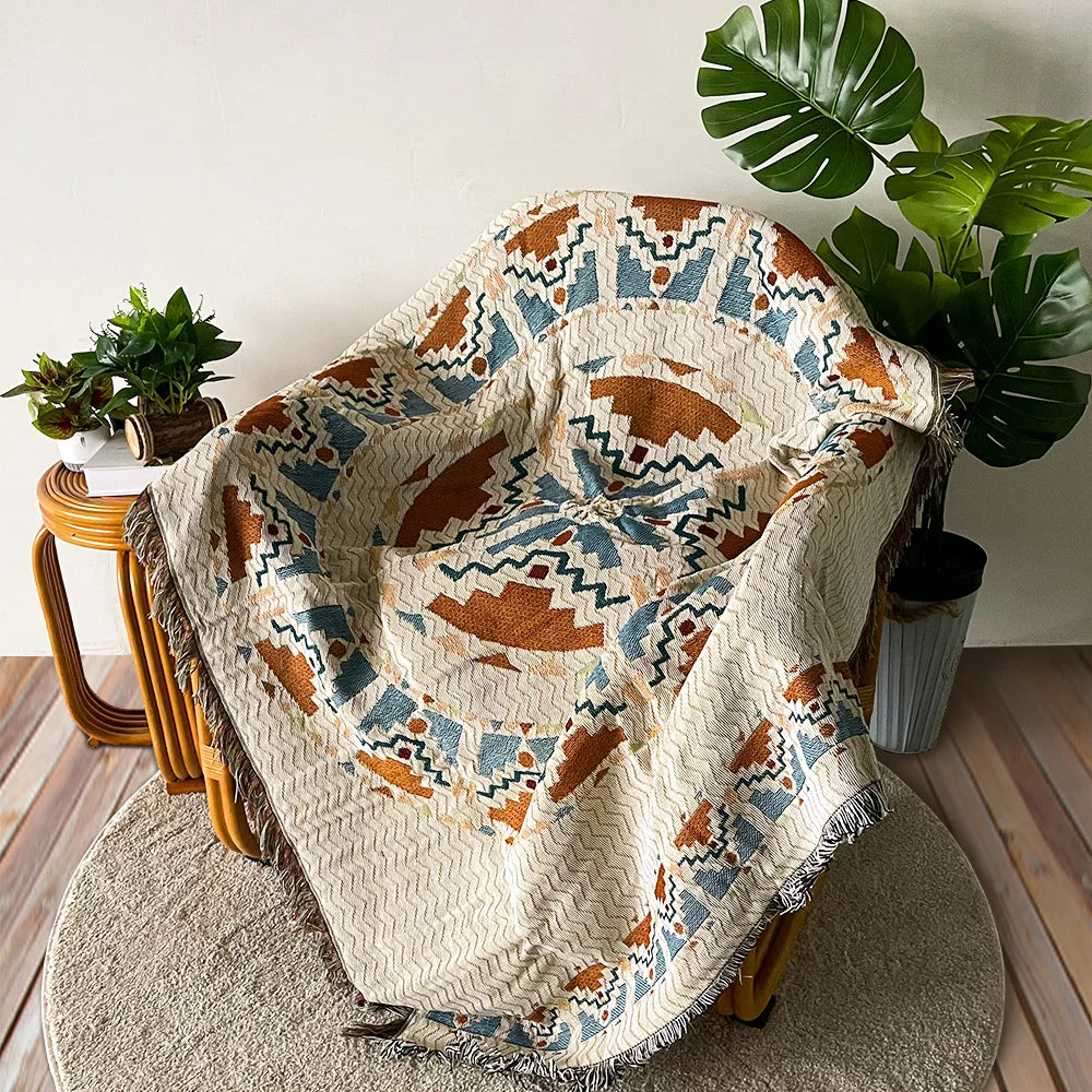A bohemian jacquard woven throw blanket draped over a wooden chair next to a large green monstera plant.