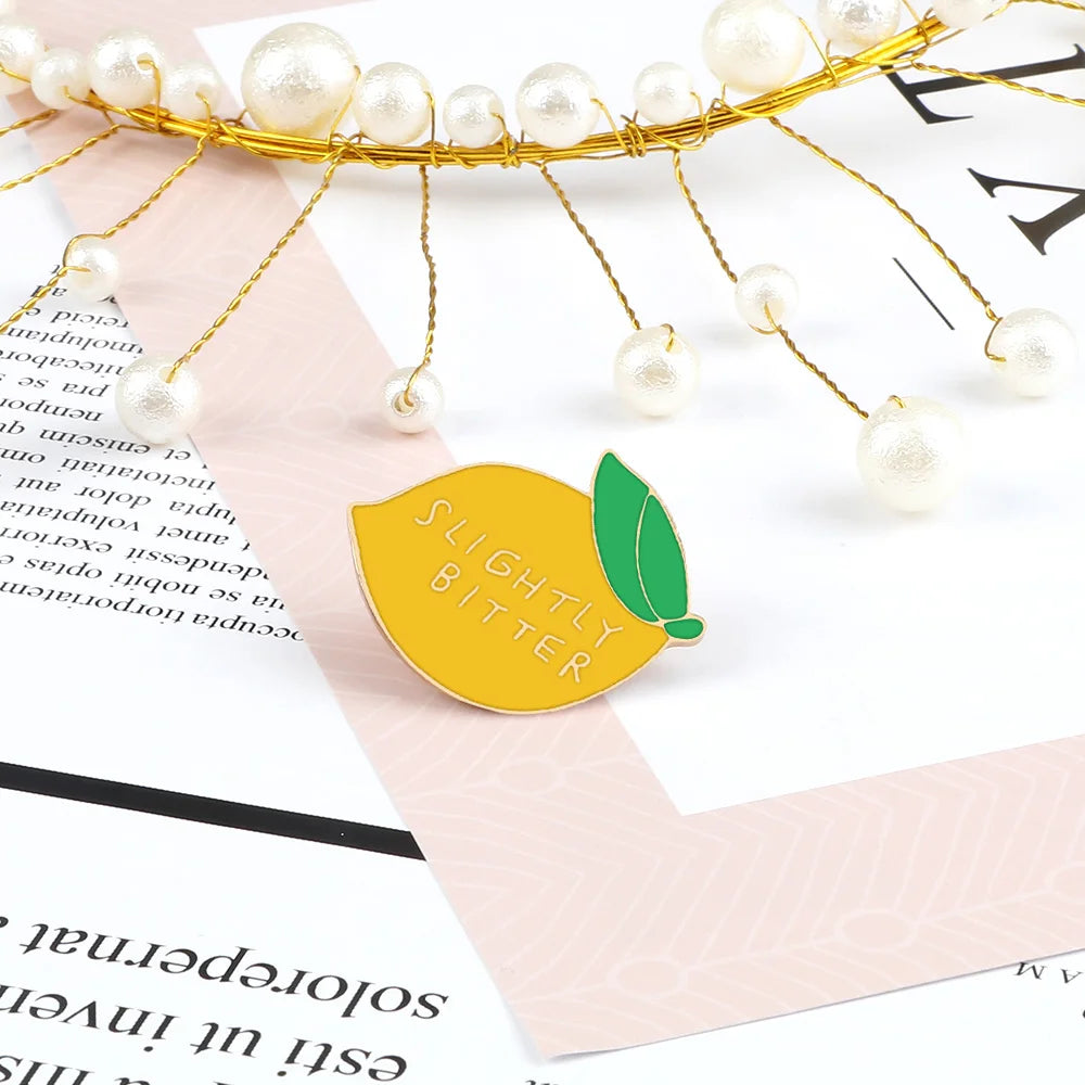 A gold wire fruit brooch pin with pearl-like beads is displayed on a pink paper background.