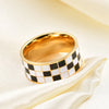 A chunky gold ring with a white and black checkerboard enamel design worn on a cream fabric.