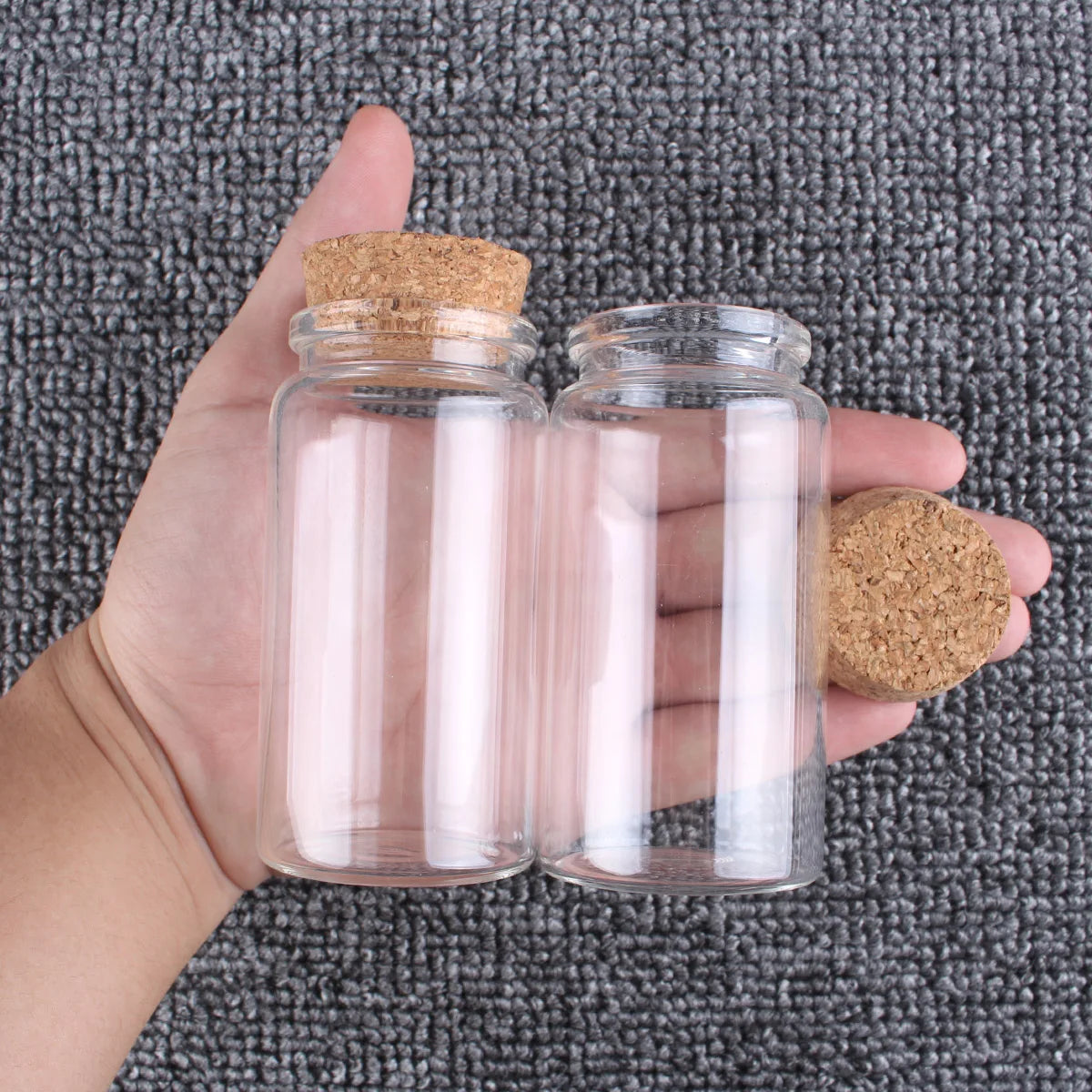 Una mano sostiene dos frascos de vidrio transparentes con tapones de corcho sobre una superficie texturizada gris.