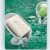 A bar of white soap is dripping into a green, textured water surface in a light gray dish.