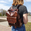 A woman is shown from the back wearing a brown PU leather fashion backpack and blue jeans outdoors.