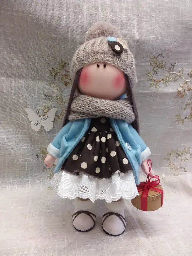 A handmade doll dressed in a gray knitted hat and blue coat with a polka dot skirt