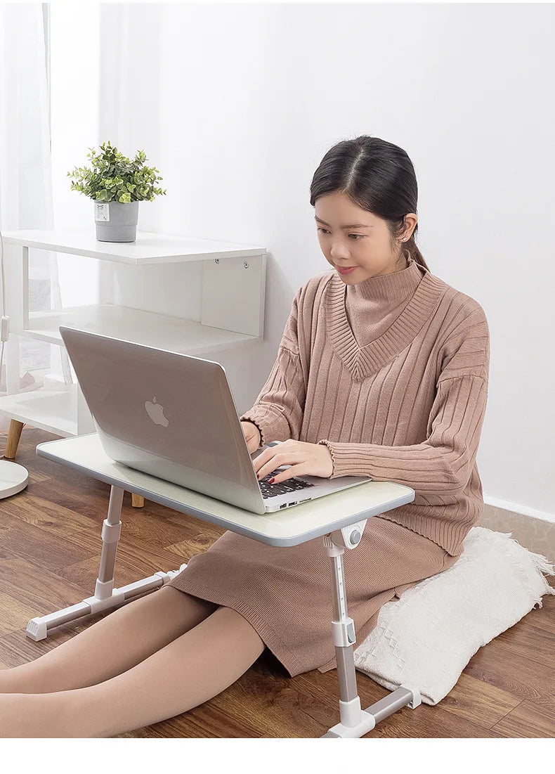 A woman is using an adjustable foldable laptop desk while lying down on a wooden floor.
