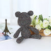 A small, dark gray metallic yarn teddy bear is displayed next to white roses and a book.
