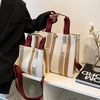 A beige and brown striped canvas crossbody tote bag with adjustable leather straps displayed on a white shelf.