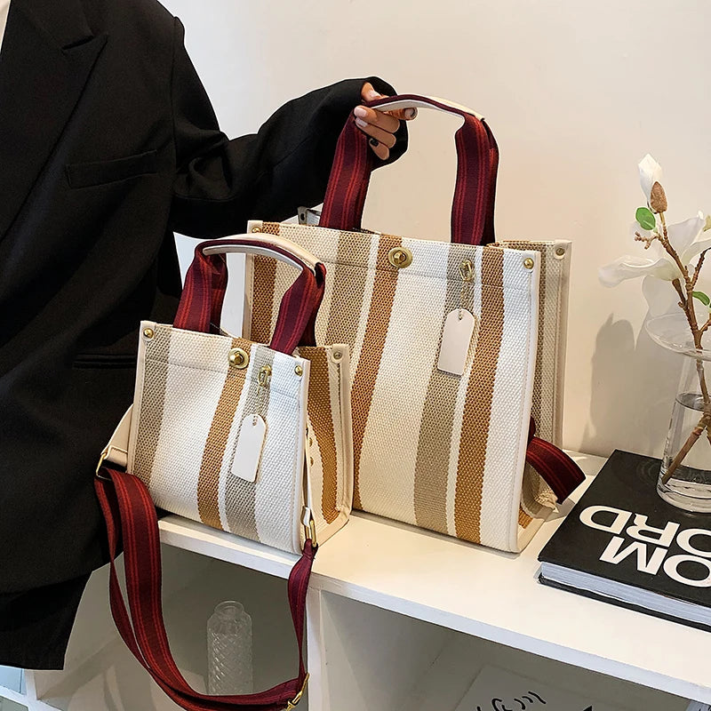 A beige and brown striped canvas crossbody tote bag with adjustable leather straps displayed on a white shelf.