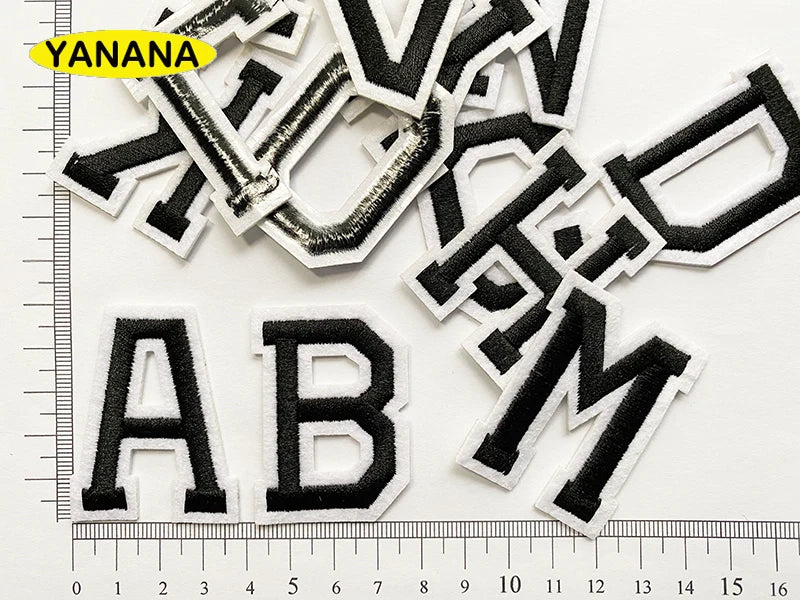 A collection of black embroidered alphabet patches, including YANANA and ABM, laid out on a white background with a ruler.