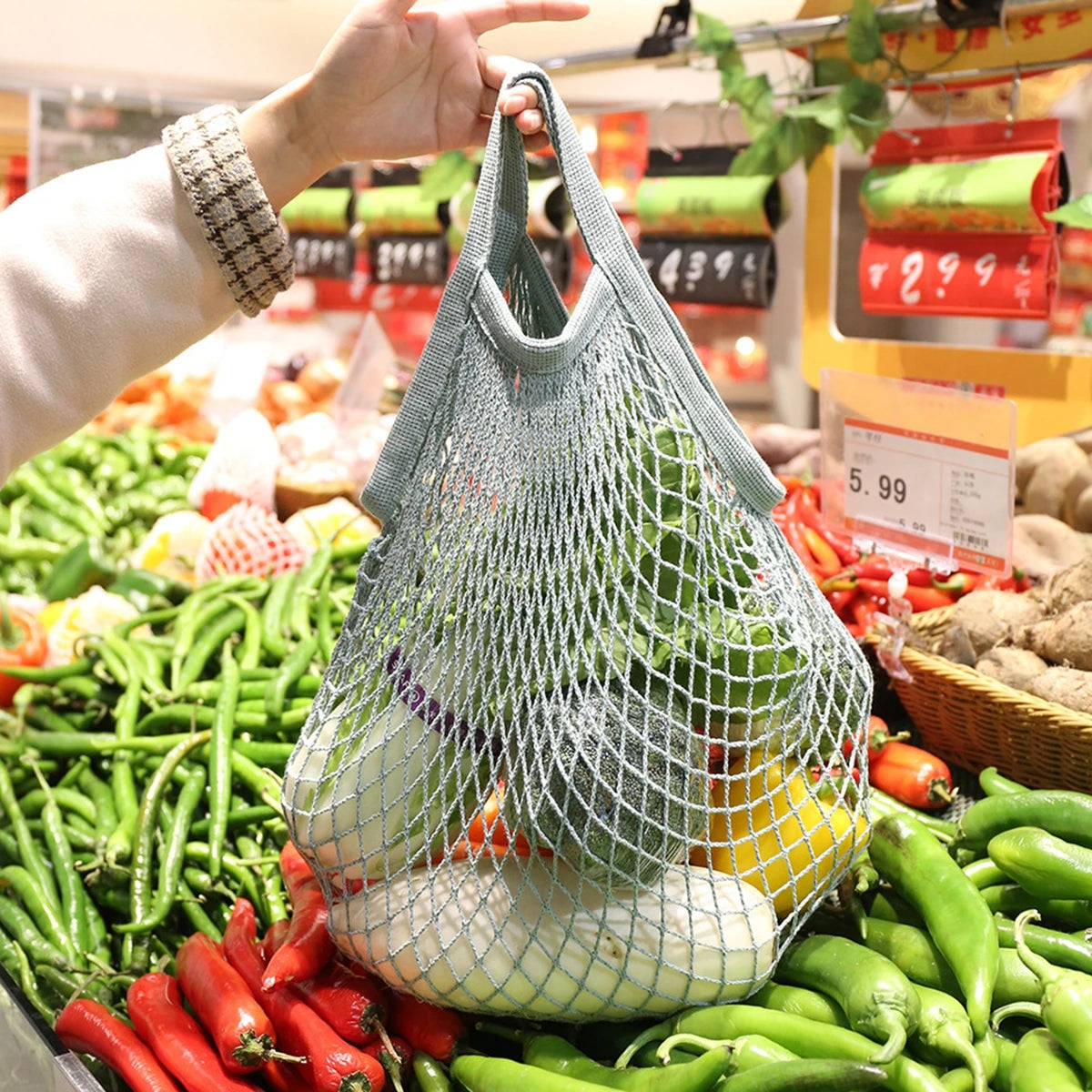 A hand is holding a mesh produce bag filled with fresh vegetables, including green peppers and tomatoes, in a market setting.