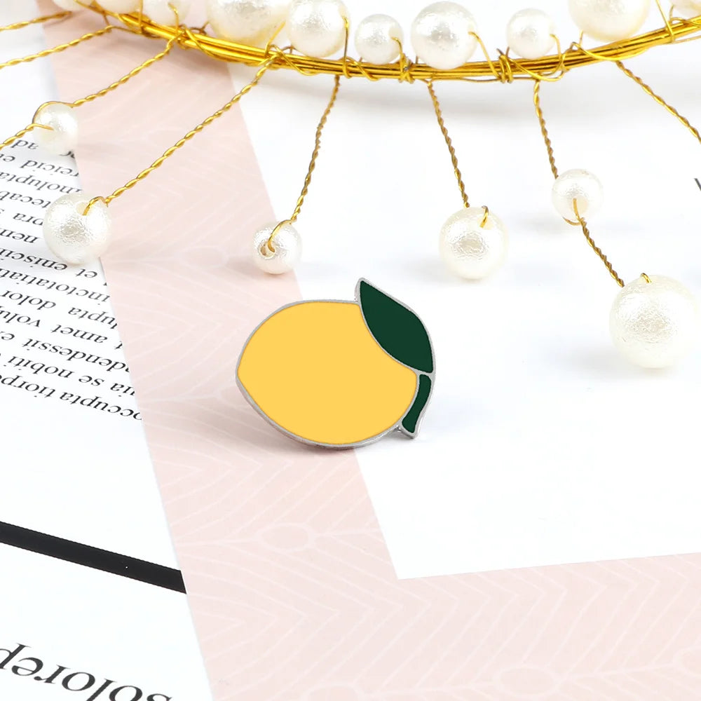 A gold wire fruit brooch pin featuring a yellow citrus fruit and green leaf, suspended by pearl-like beads.