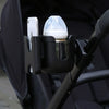 A black stroller cup and phone holder device is mounted on a stroller handlebar, featuring a clear glass cup and a smartphone