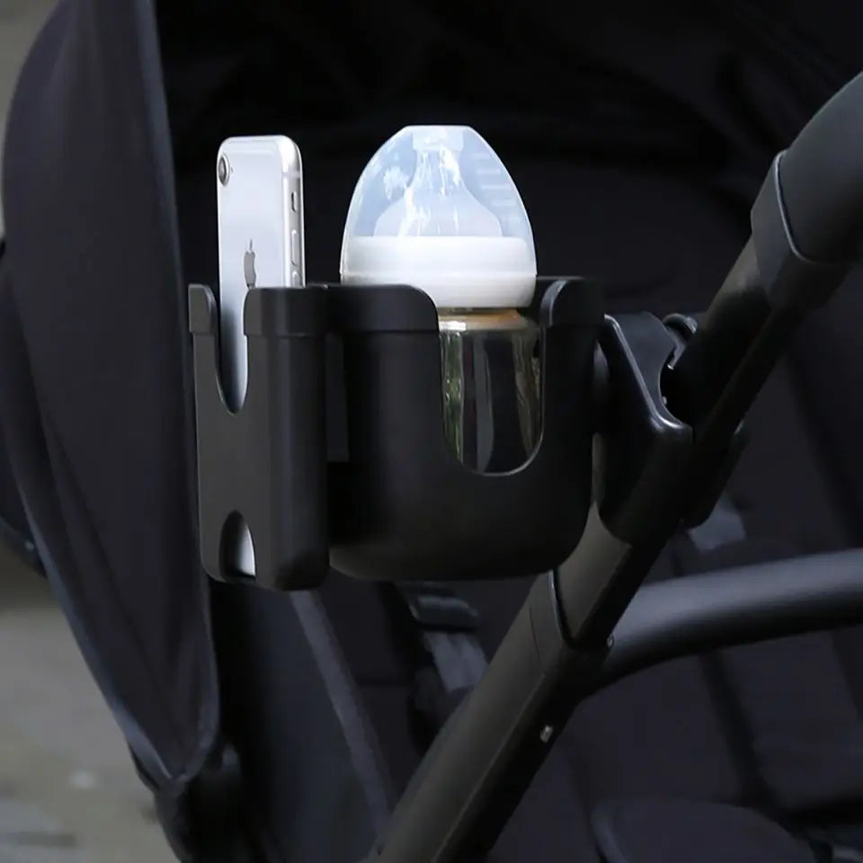 A black stroller cup and phone holder device is mounted on a stroller handlebar, featuring a clear glass cup and a smartphone