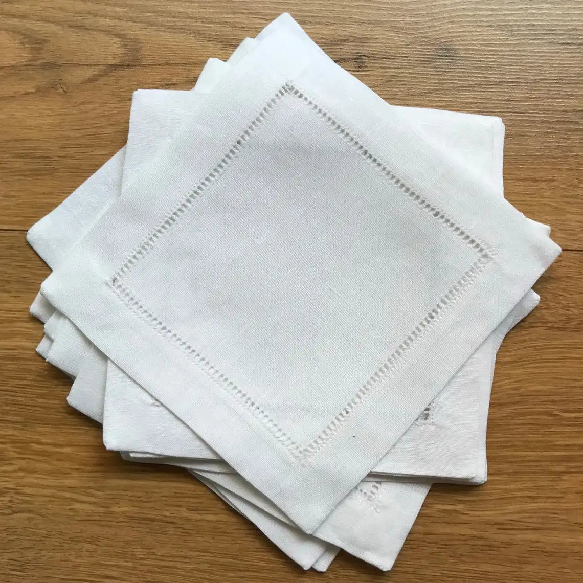 A stack of six white linen hemstitch napkins folded neatly on a wooden surface.