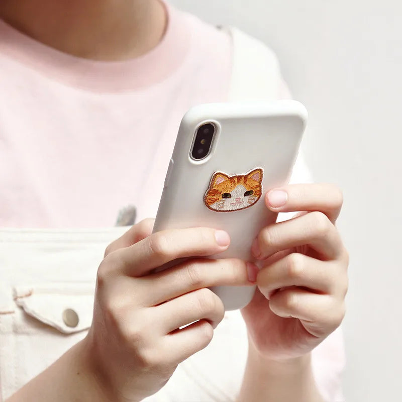 A person holds a white smartphone displaying an embroidered orange cat iron-on patch.