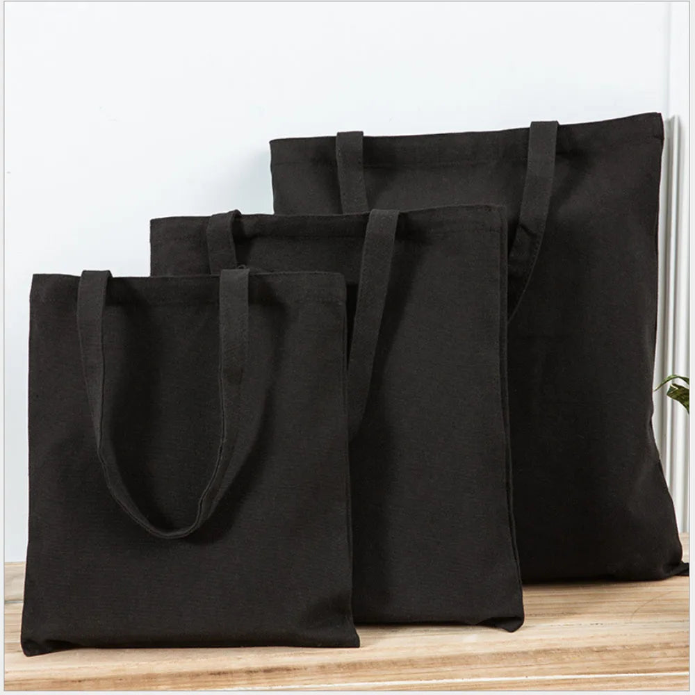 A stack of black canvas tote bags with handles resting on a wooden surface.