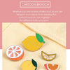 A collection of enamel fruit brooches featuring citrus slices like lemon, orange, and lime on a beige fabric background.