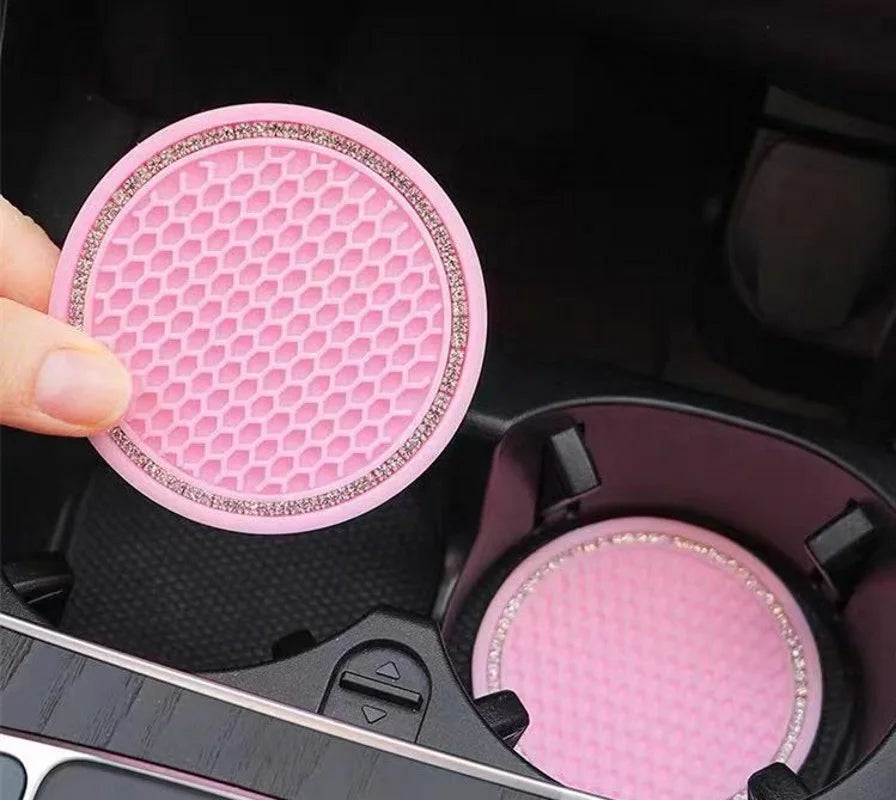 A hand holds a pink, rhinestone-encrusted non-slip cup mat for cars inside a black storage compartment.