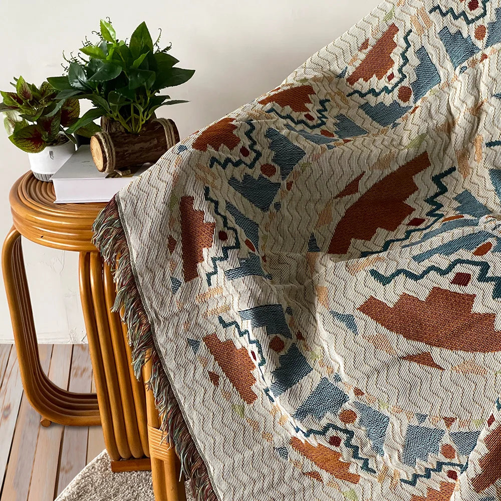 A woven throw blanket featuring a bohemian jacquard pattern in shades of rust, blue, and beige draped over a wooden stool.
