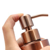 A hand is holding a rose gold stainless steel liquid soap dispenser pump bottle.