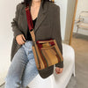 A woman wearing a brown blazer and jeans holds a canvas crossbody tote bag with an adjustable strap.
