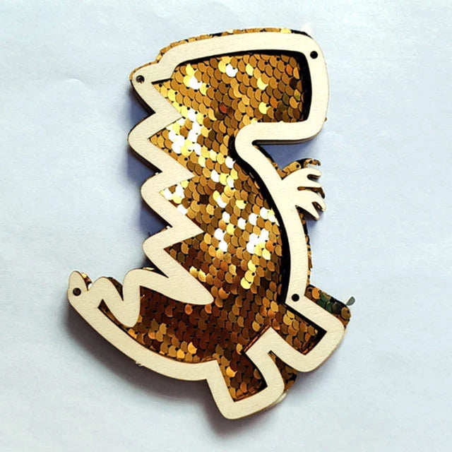 A wooden busy board piece shaped like a dinosaur, covered in gold and silver sequins.