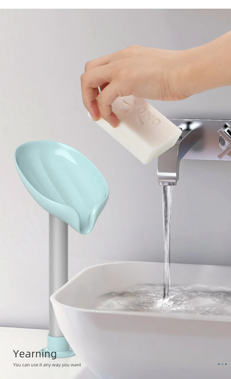 A hand is placing a white leaf-shaped suction cup soap onto a chrome faucet, with water flowing into a white sink.