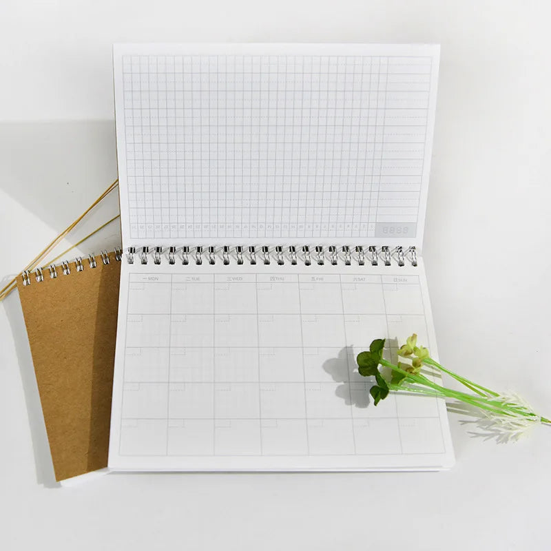 An open A5 spiral-bound monthly planner with ruled pages and a brown cover resting on a white surface.