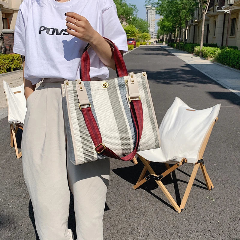 A person wearing light pants holds a beige and striped canvas crossbody tote bag with a red strap.